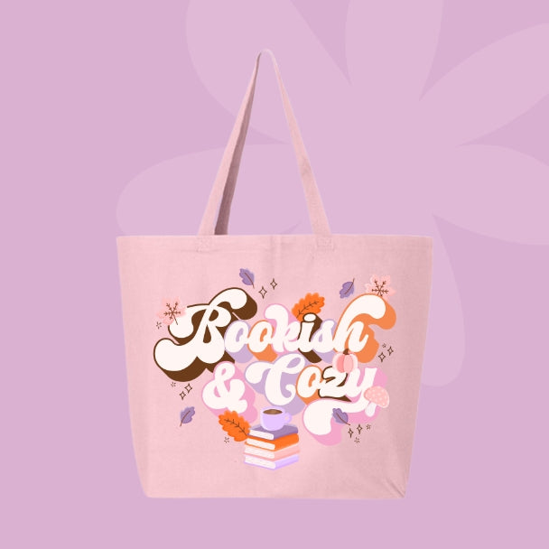 Pink tote bag with bold retro lettering that says “Bookish & Cozy” surrounded by fall leaves, mushrooms, and stacked books.