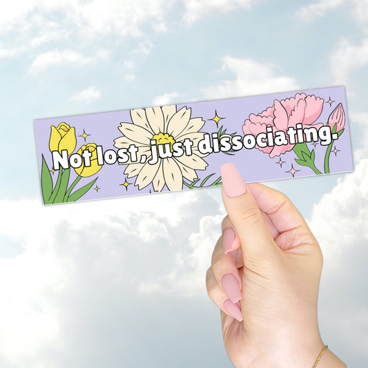 A hand holding a pastel purple bumper sticker that reads "Not lost, just dissociating" with illustrated flowers and sparkles.