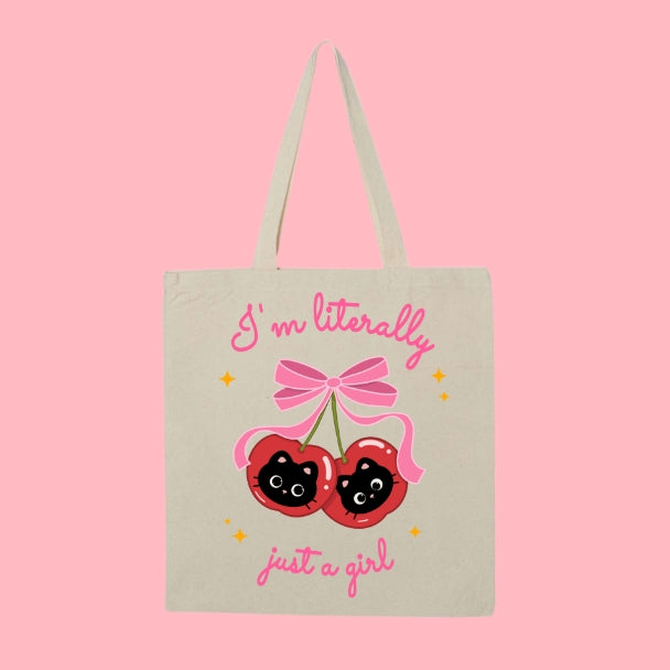 Beige tote bag with a design of two cherries featuring black cat faces and a pink bow, with text that says “I’m Literally Just a Girl.”