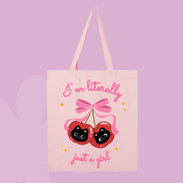 Pink tote bag with a design of two cherries featuring black cat faces and a pink bow, with text that says “I’m Literally Just a Girl.”