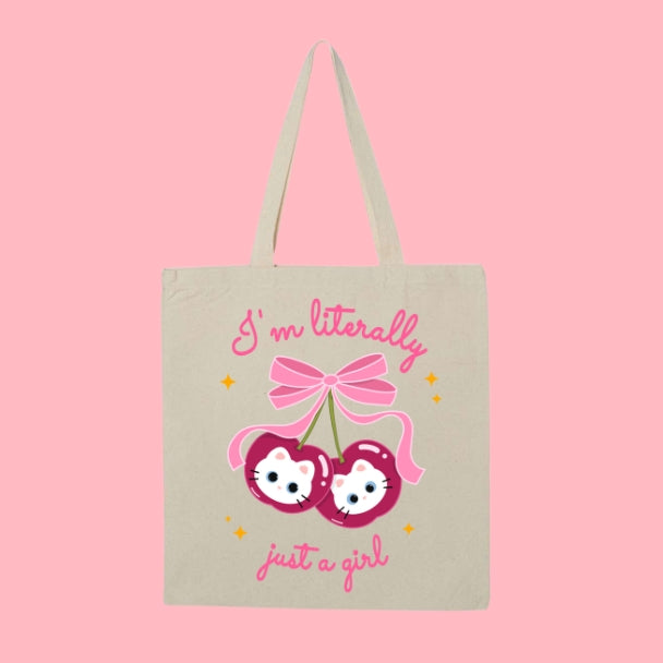 Beige tote bag with a design of two cherries featuring white cat faces and a pink bow, with text that says “I’m Literally Just a Girl.”