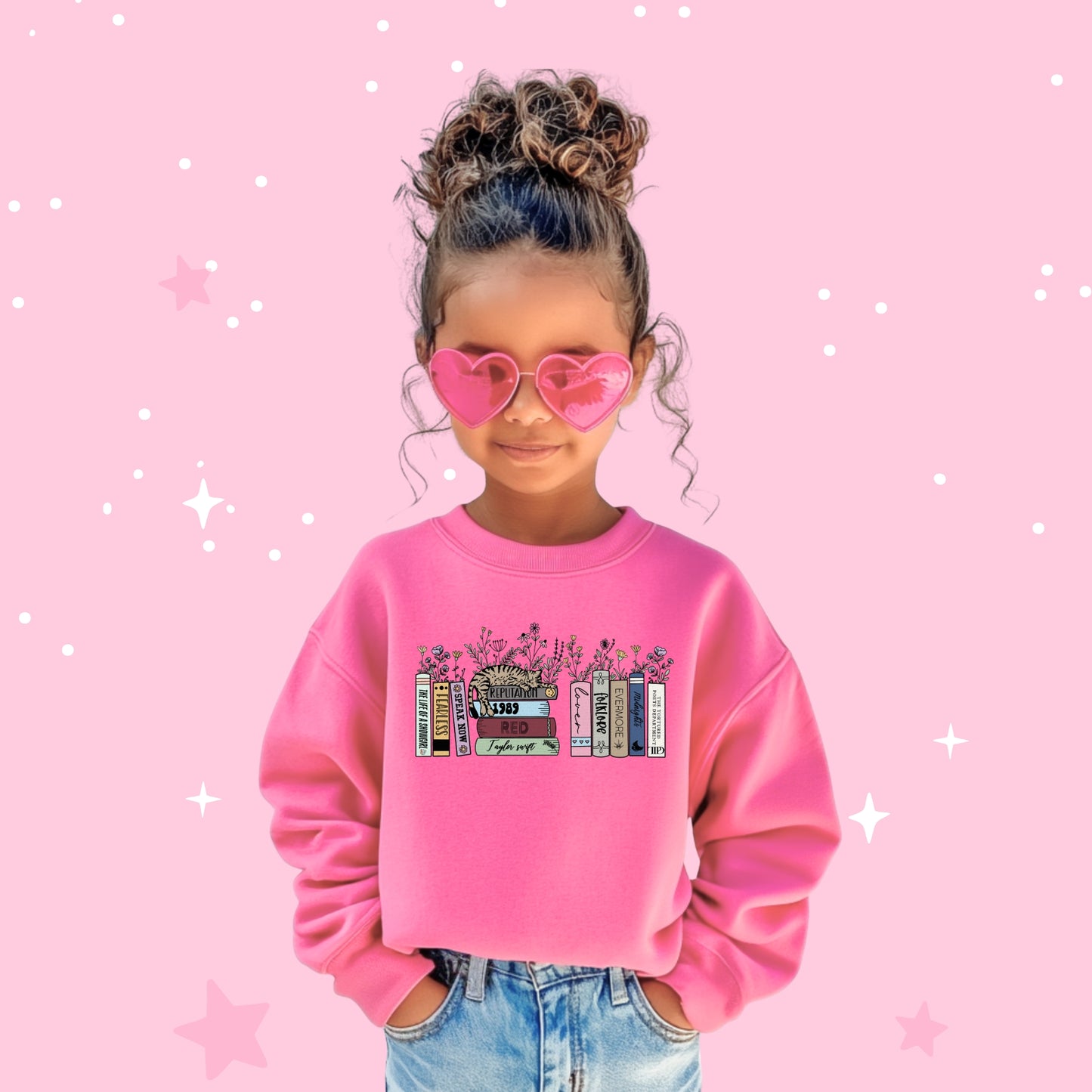 A young girl wearing a pink Taylor Swift-themed sweatshirt with illustrated books representing each of Taylor Swift’s eras from Debut to The Life of a Showgirl, topped with a cat and surrounded by flowers, against a soft pink starry background.