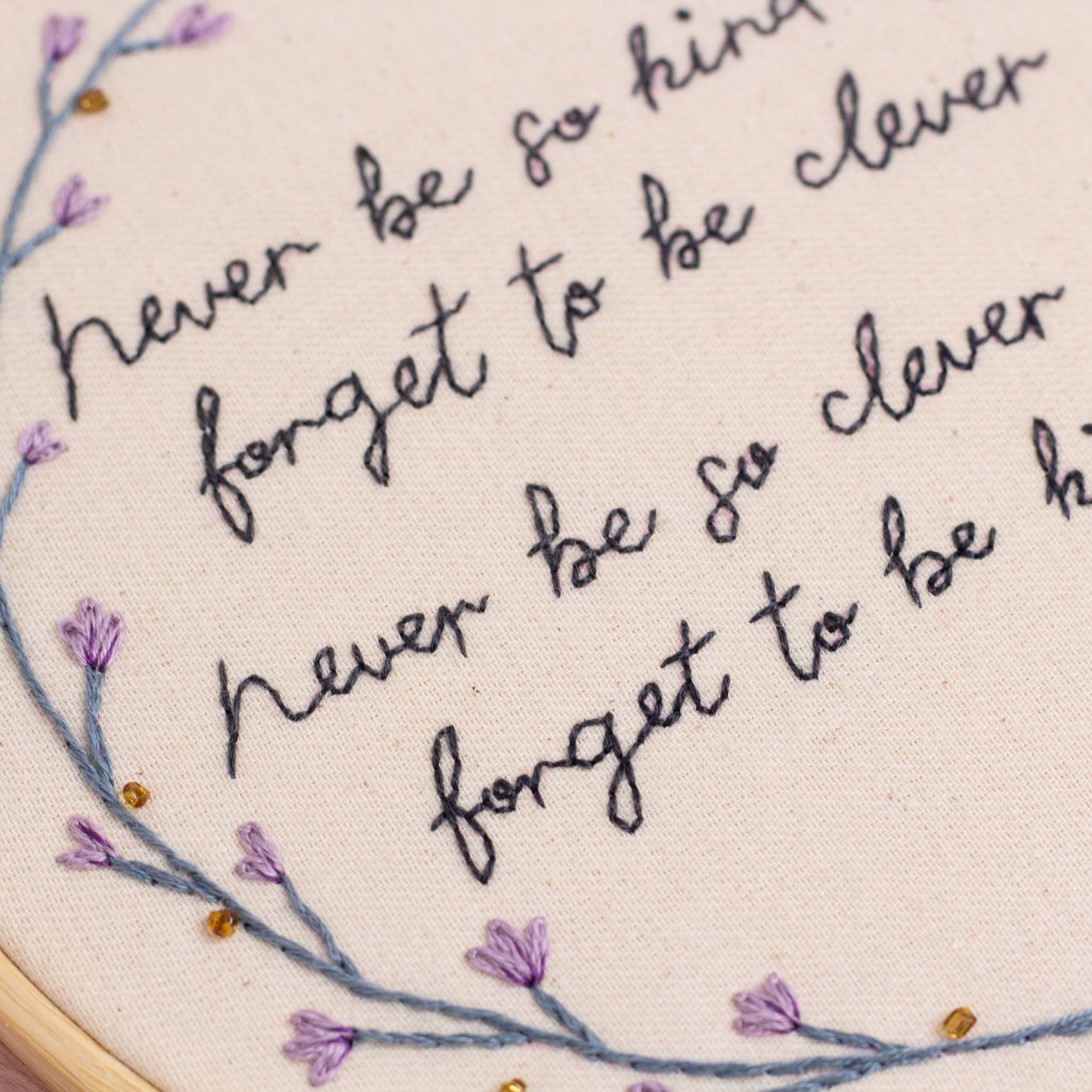 Close up of hand embroidered never be so kind quote with purple floral embroidery on cotton fabric
