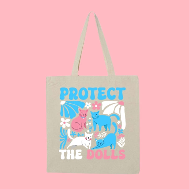 Beige tote bag with bold text “Protect the Dolls” and colorful illustrations of pink, blue, and white cats surrounded by flowers.