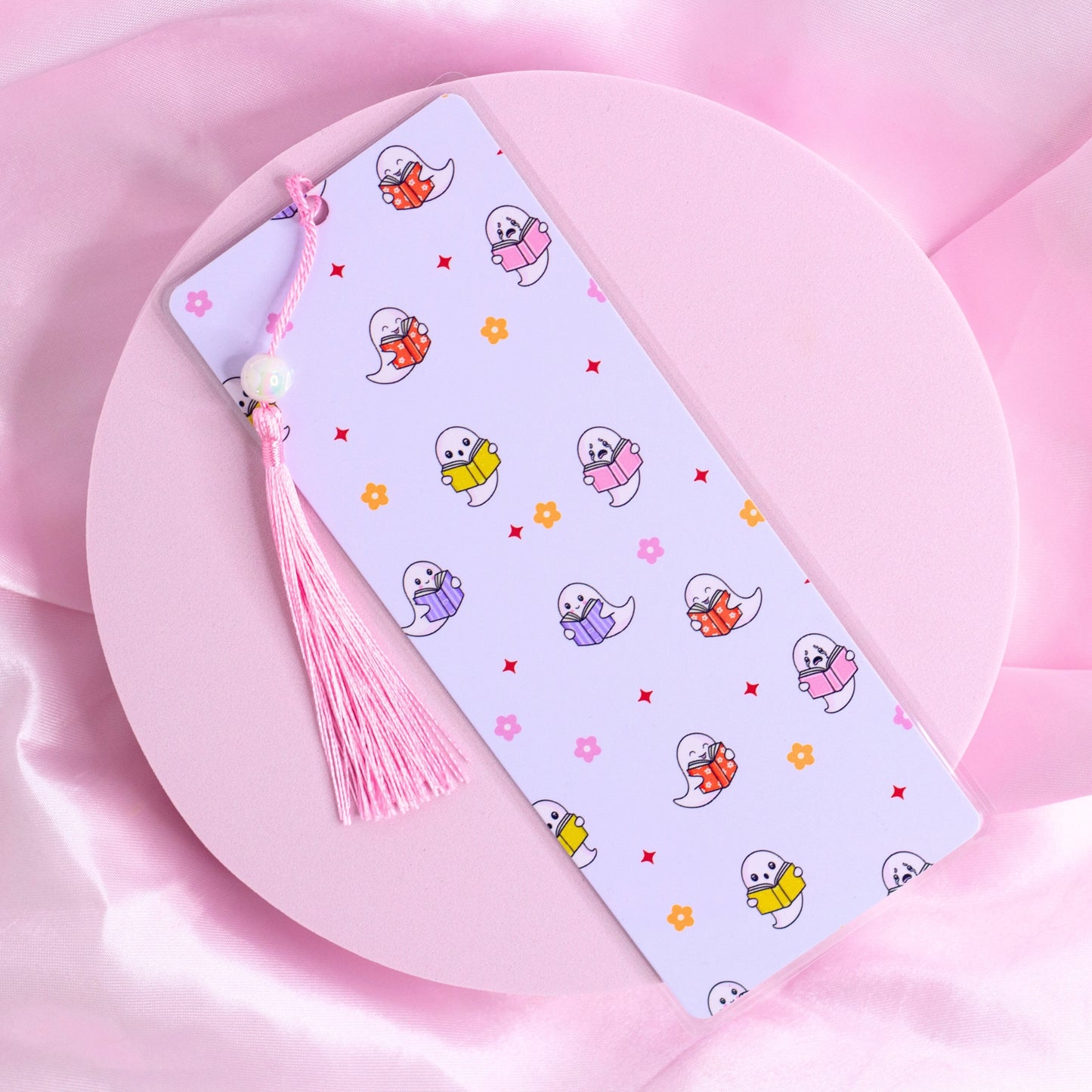 Lilac bookmark with colorful pastel ghosts holding books, surrounded by flowers and stars, with a pink tassel.
