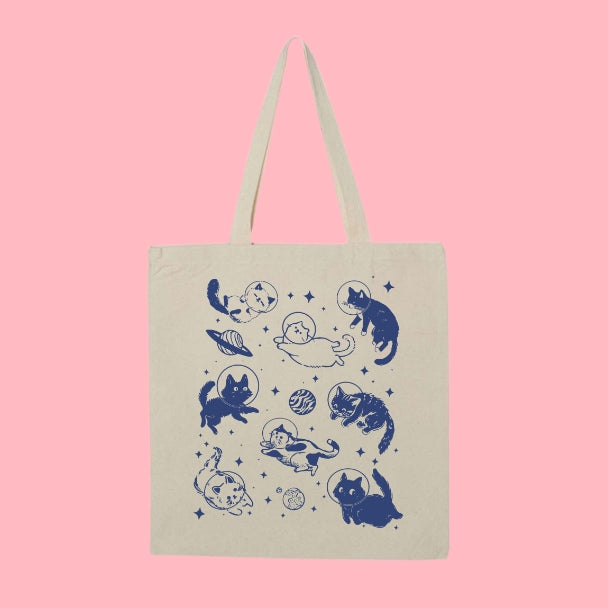 Beige tote bag with a navy design of cats floating in space with planets, stars, and astronaut helmets.