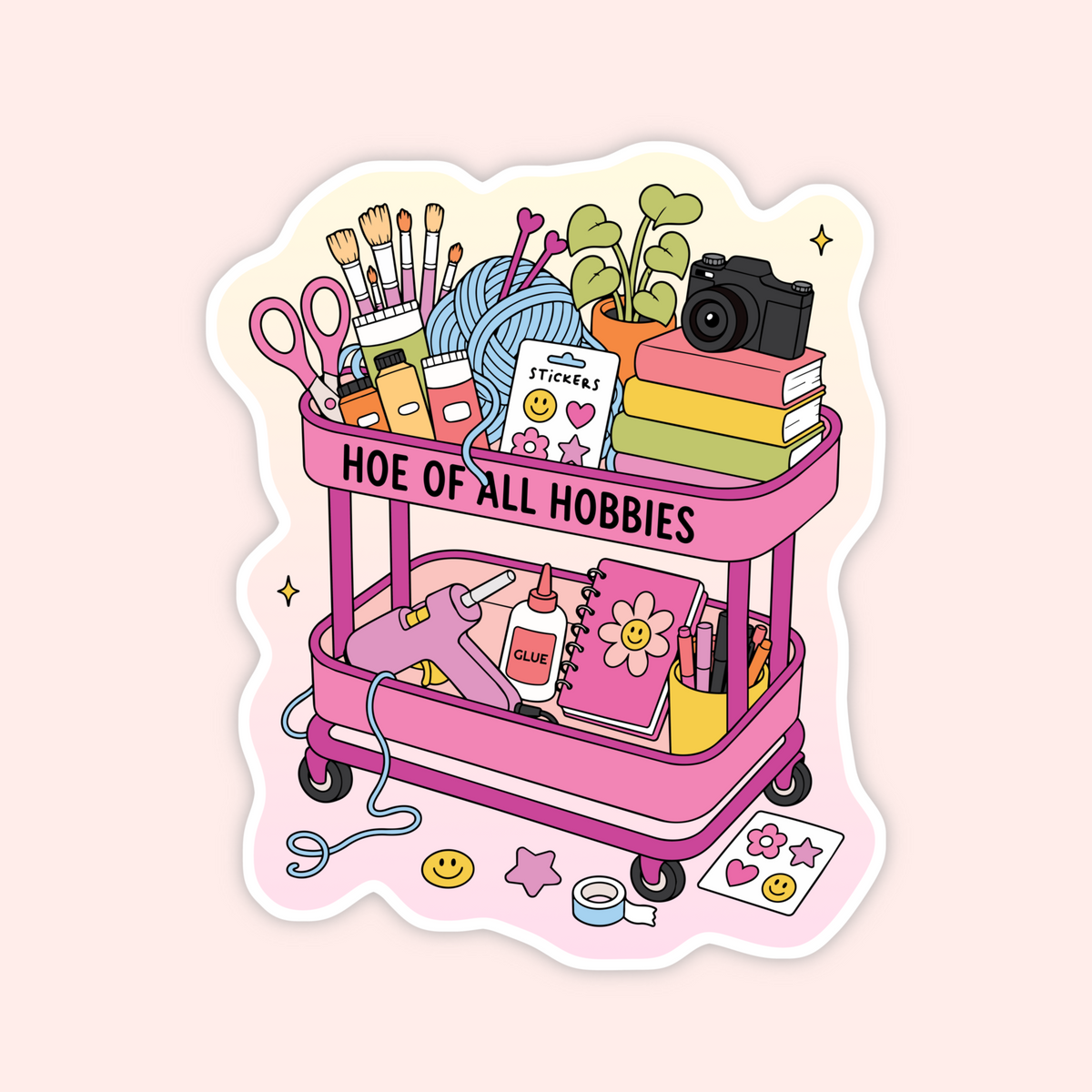 A die-cut sticker featuring a pink rolling cart filled with colorful hobby and craft supplies, including scissors, paintbrushes, yarn, books, a camera, a hot air balloon, a notebook, and stickers. The cart has the text 'HOE OF ALL HOBBIES' on the top rail.