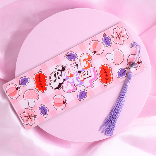 Pink gingham bookmark with retro text “Bookish & Cozy” surrounded by leaves, mushrooms, and pumpkins, with purple tassel.