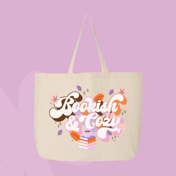 Beige tote bag with bold retro lettering that says “Bookish & Cozy” surrounded by fall leaves, mushrooms, and stacked books.