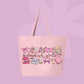 Large pink canvas tote bag featuring the words bookish girlie surrounded by books, bows, coffee cups, flowers, a cozy chair, a cat, and reading-inspired doodles.