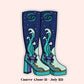 A stylized illustration of a pair of tall knee-high boots with a Cancer zodiac theme, featuring a navy blue upper decorated with a teal Cancer symbol and stars, and a light teal lower section with a purple crab and wavy teal details. The boots are shown on a pale pink background with the text 'Cancer (June 21 - July 22)' underneath.