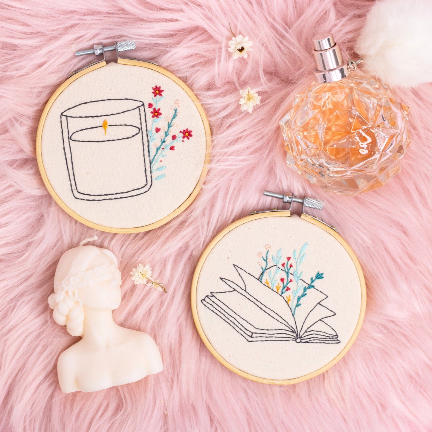 hand embroidered candle and open book wall art hoops on pink background with flowers and perfume styling