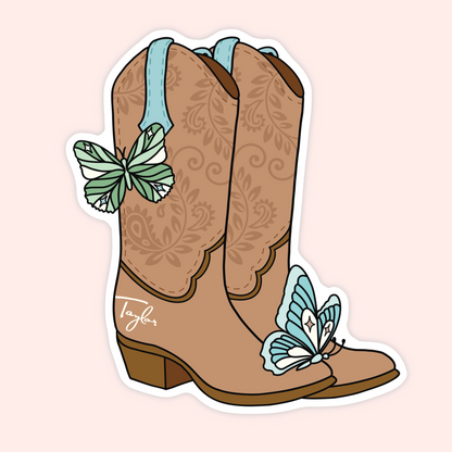 A die-cut sticker featuring an illustration of a pair of tan western cowboy boots with floral scrollwork, light blue pull tabs, and two light blue butterflies, shown on a pale pink background.
