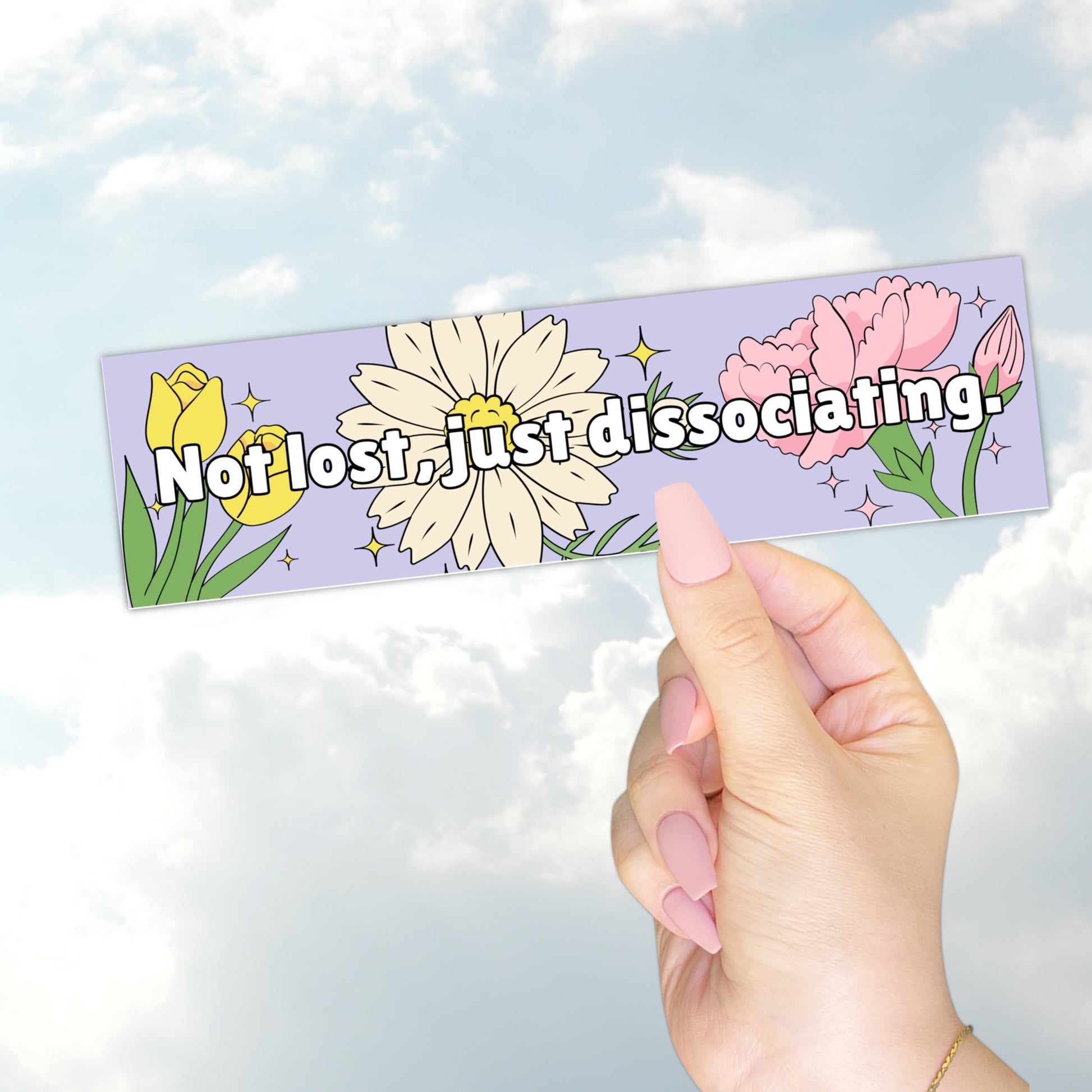 A hand holding a pastel purple bumper sticker that reads "Not lost, just dissociating" with illustrated flowers and sparkles.