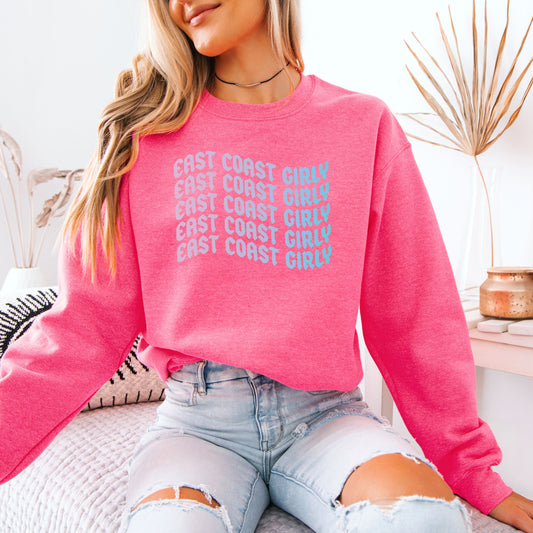 A woman wearing a bright pink sweatshirt with stacked blue text reading “East Coast Girly.” She’s sitting casually on a bed in ripped jeans, giving off relaxed coastal girl vibes with natural lighting and a modern neutral background.