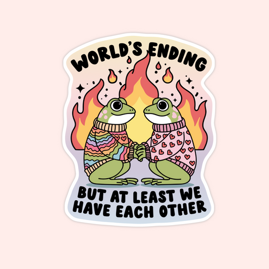 Two green frogs in cozy rainbow and heart sweaters holding hands with flames in the background. Text says “World’s Ending But At Least We Have Each Other.”