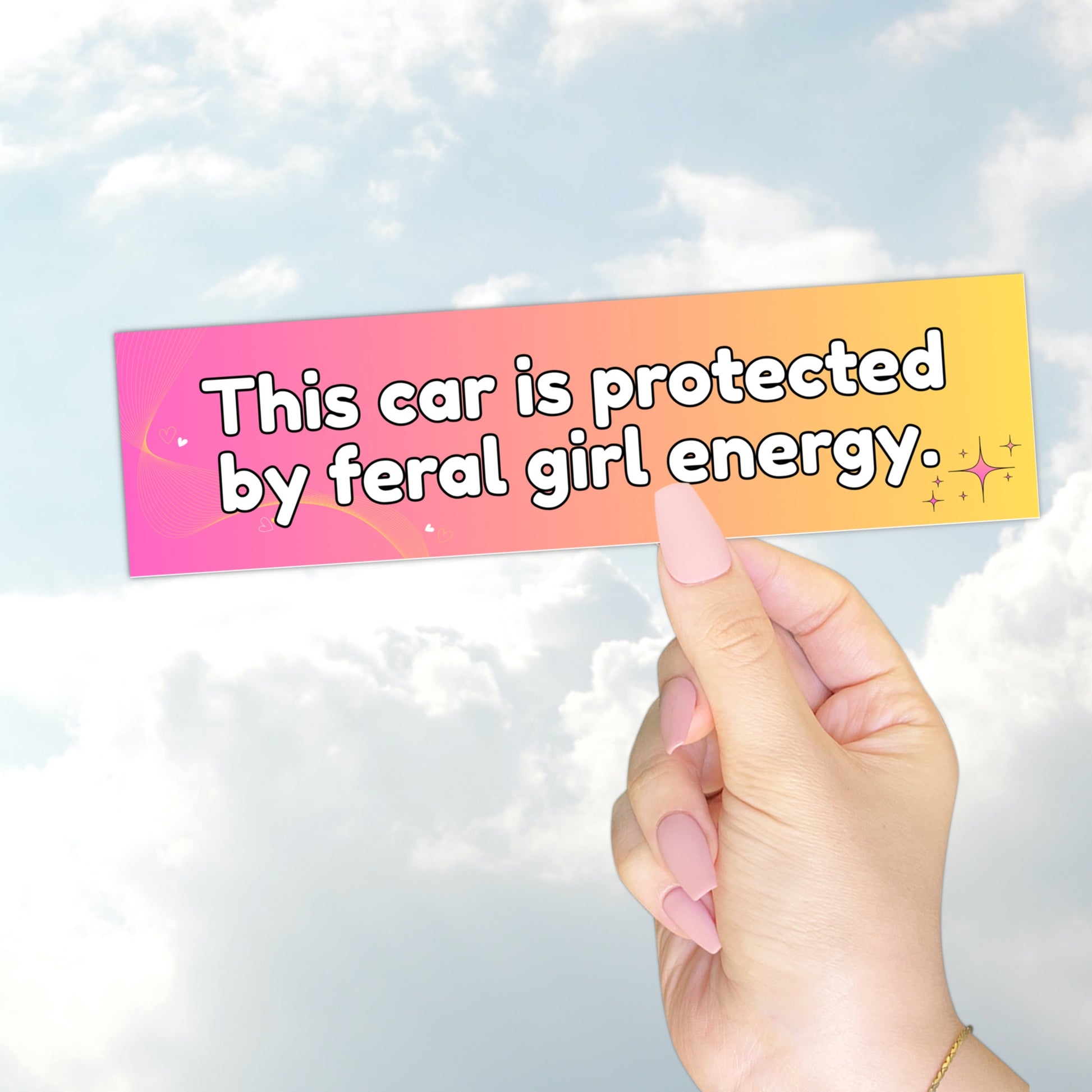 A hand holding a gradient pink-to-yellow bumper sticker with the text "This car is protected by feral girl energy" and sparkles.