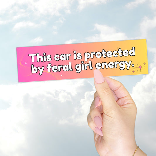 A hand holding a gradient pink-to-yellow bumper sticker with the text "This car is protected by feral girl energy" and sparkles.