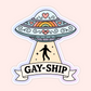 A pastel UFO with rainbow and heart details beaming up a silhouette. Text below says “Gay-Ship.”