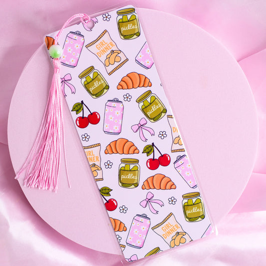 Pink bookmark with illustrations of pickles, croissants, cherries, bows, and snack bags with a pink tassel.