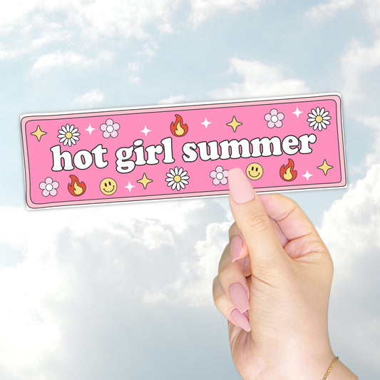 A hand holding a pink bumper sticker that says "hot girl summer" surrounded by daisies, flames, smiley faces, and sparkles.