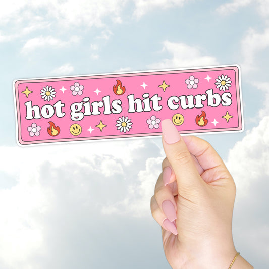 A hand holding a pink bumper sticker that says "hot girls hit curbs" surrounded by daisies, flames, smiley faces, and sparkles.