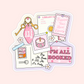 A pink bookish collage featuring a motel keychain, library card, candle, postcards, and the phrase “I’m All Booked.”