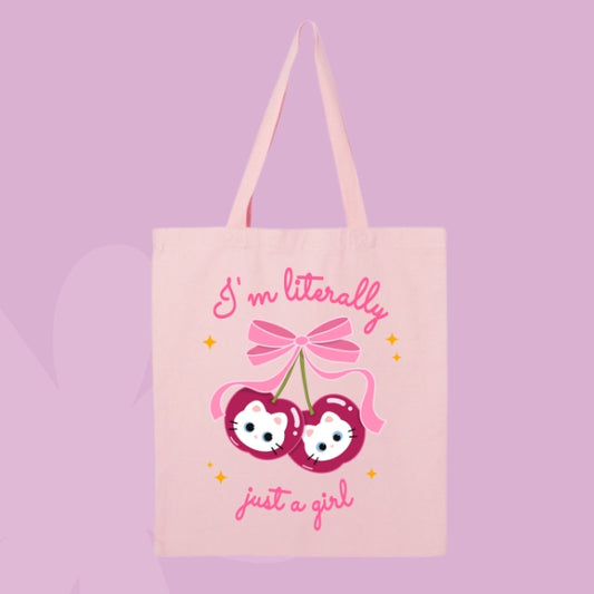 Pink tote bag with a design of two cherries featuring white cat faces and a pink bow, with text that says “I’m Literally Just a Girl.”