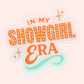 Retro-inspired sticker with bold orange lettering reading “In My Showgirl Era” with sparkles and a soft pastel background.
