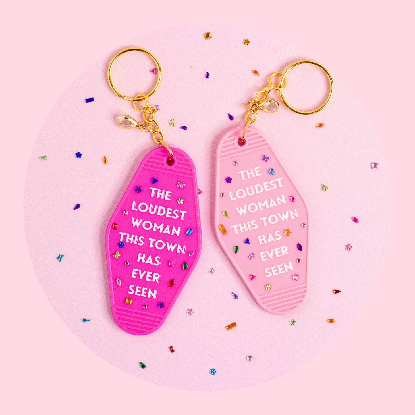 two retro motel keychains in bright pink and soft pink with bold text and colorful rhinestone details styled on pink surface