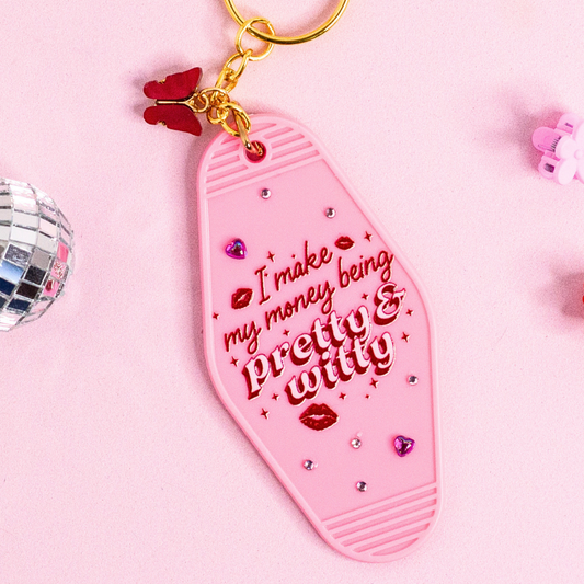 close up of pink retro motel keychain with red lettering “pretty & witty”, rhinestone details and glossy finish with gold hardware