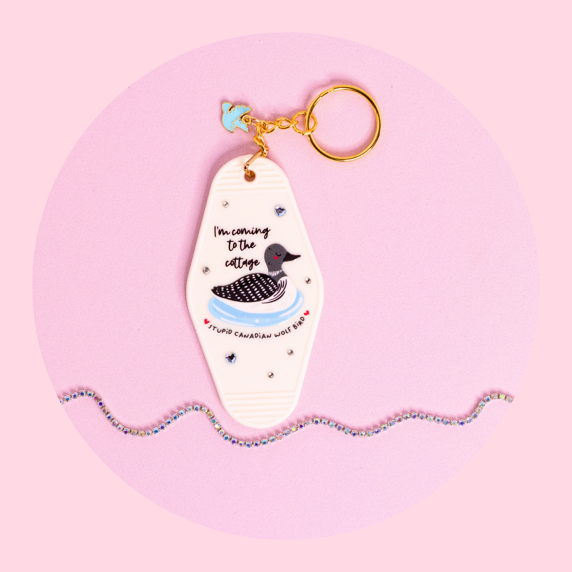 white motel style keychain with loon illustration and “i’m coming to the cottage” text, rhinestones and gold key ring on pink background