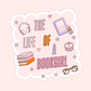 Pink sticker with the words “The Life of a Bookgirl” surrounded by books, headphones, a candle, glasses, and an e-reader.