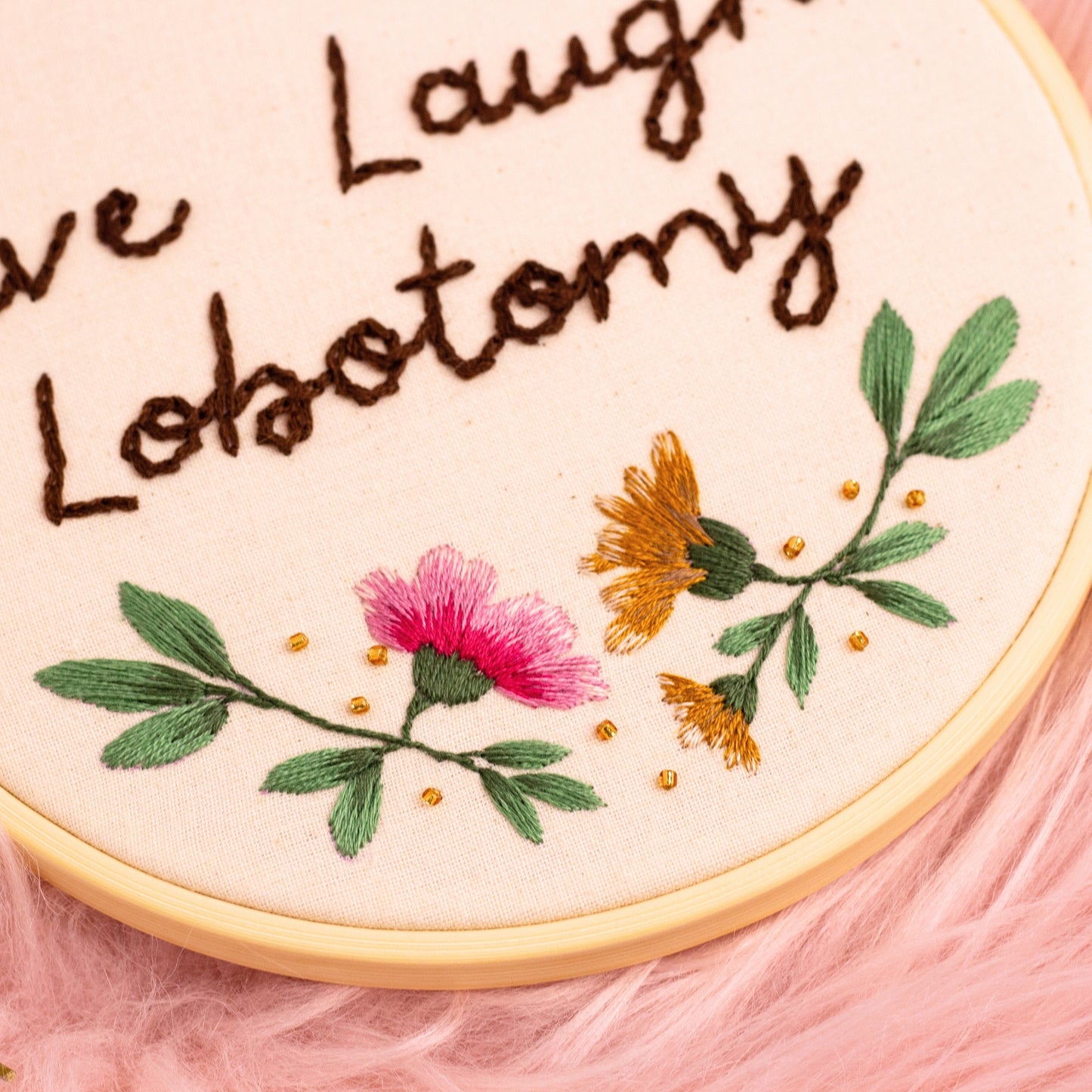 Close-up of handmade embroidery hoop featuring pink stitched flower with green leaves and gold bead accents on neutral fabric