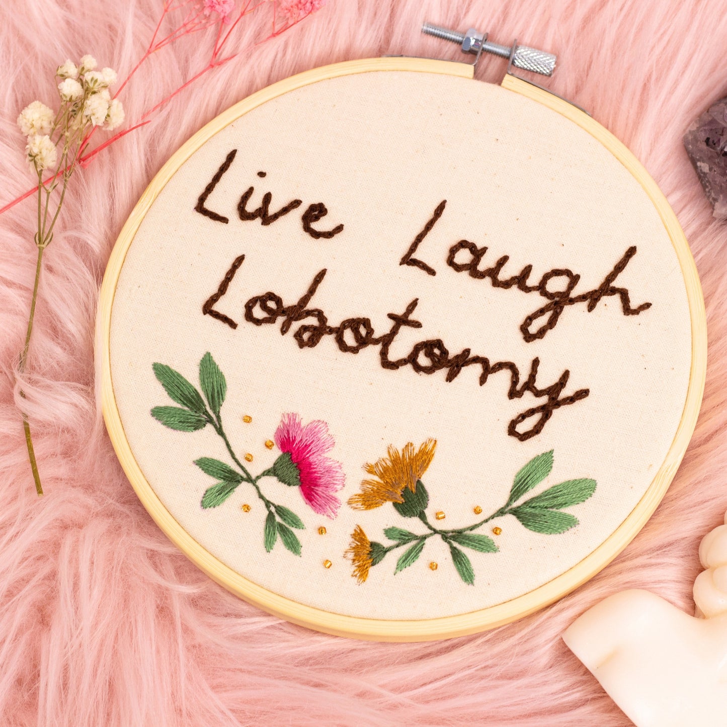Full view of Live Laugh Lobotomy embroidery hoop with floral accents on soft pink textured background