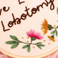 Angled close-up of embroidered floral details and Live Laugh Lobotomy text stitched in brown thread on fabric hoop