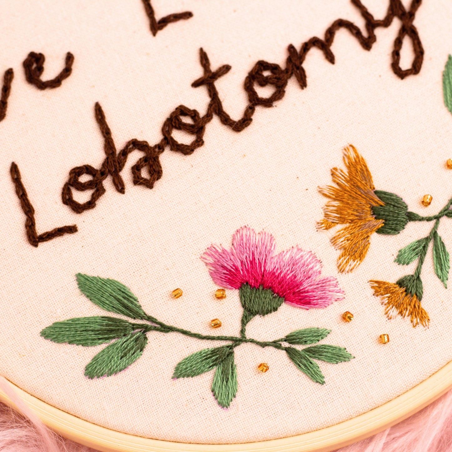 Angled close-up of embroidered floral details and Live Laugh Lobotomy text stitched in brown thread on fabric hoop