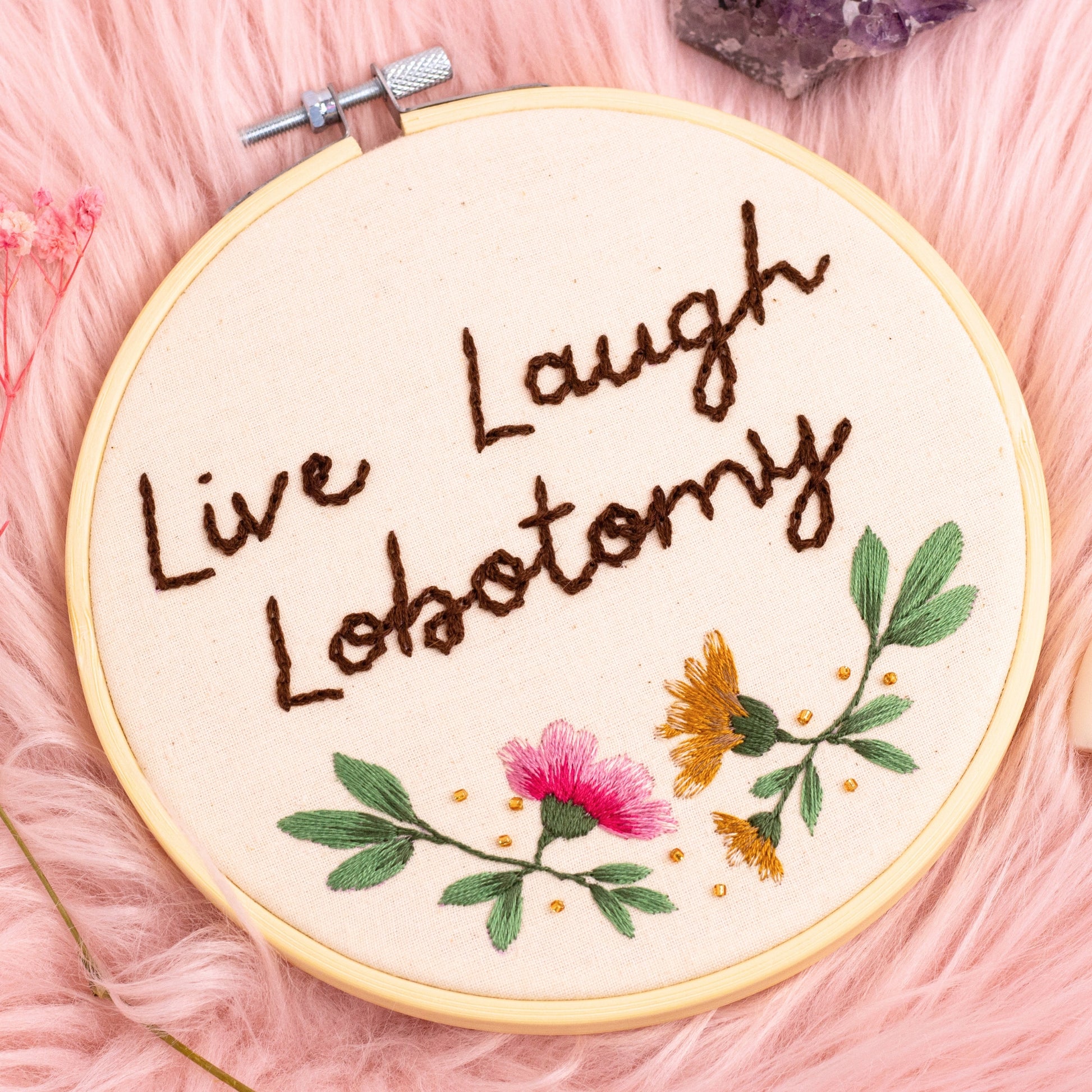 Handmade Live Laugh Lobotomy embroidery hoop with floral design displayed on pink faux fur background with crystals and dried flowers