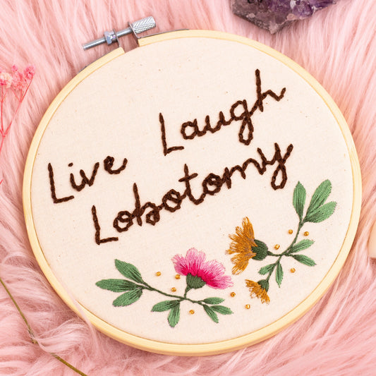 Handmade Live Laugh Lobotomy embroidery hoop with floral design displayed on pink faux fur background with crystals and dried flowers