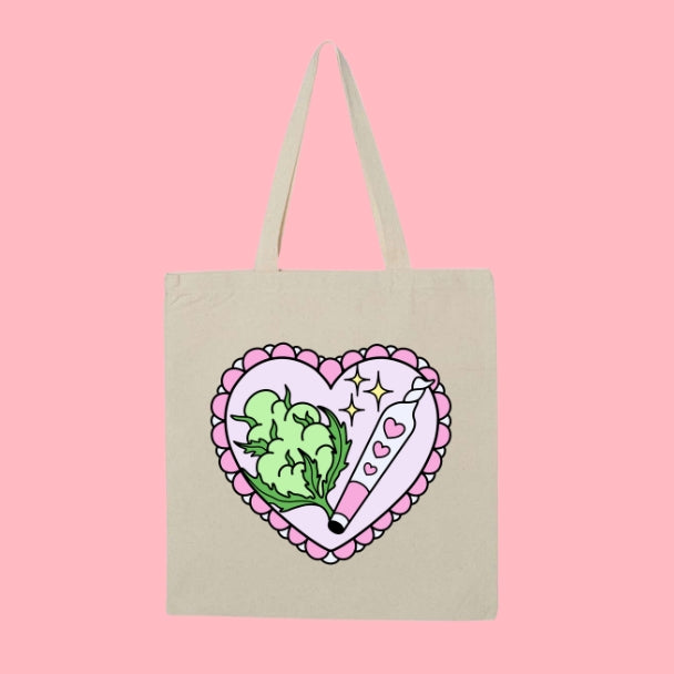 Beige tote bag with a pink heart design featuring green cannabis leaves and a sparkly rolled joint.