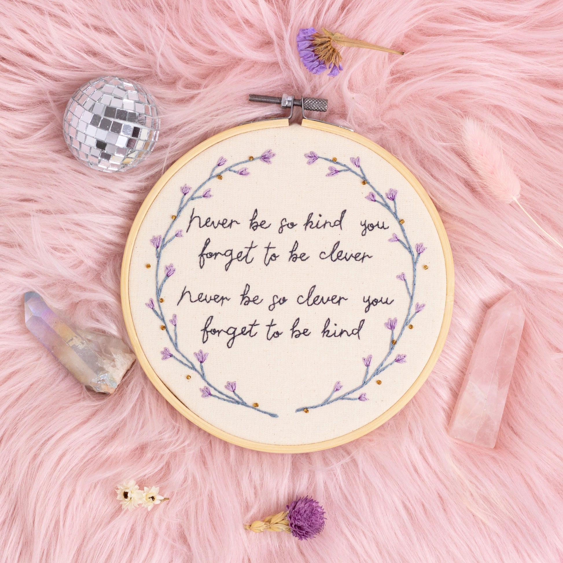 Never be so kind quote embroidery hoop wall art with purple flowers styled on pink background with crystals and disco ball