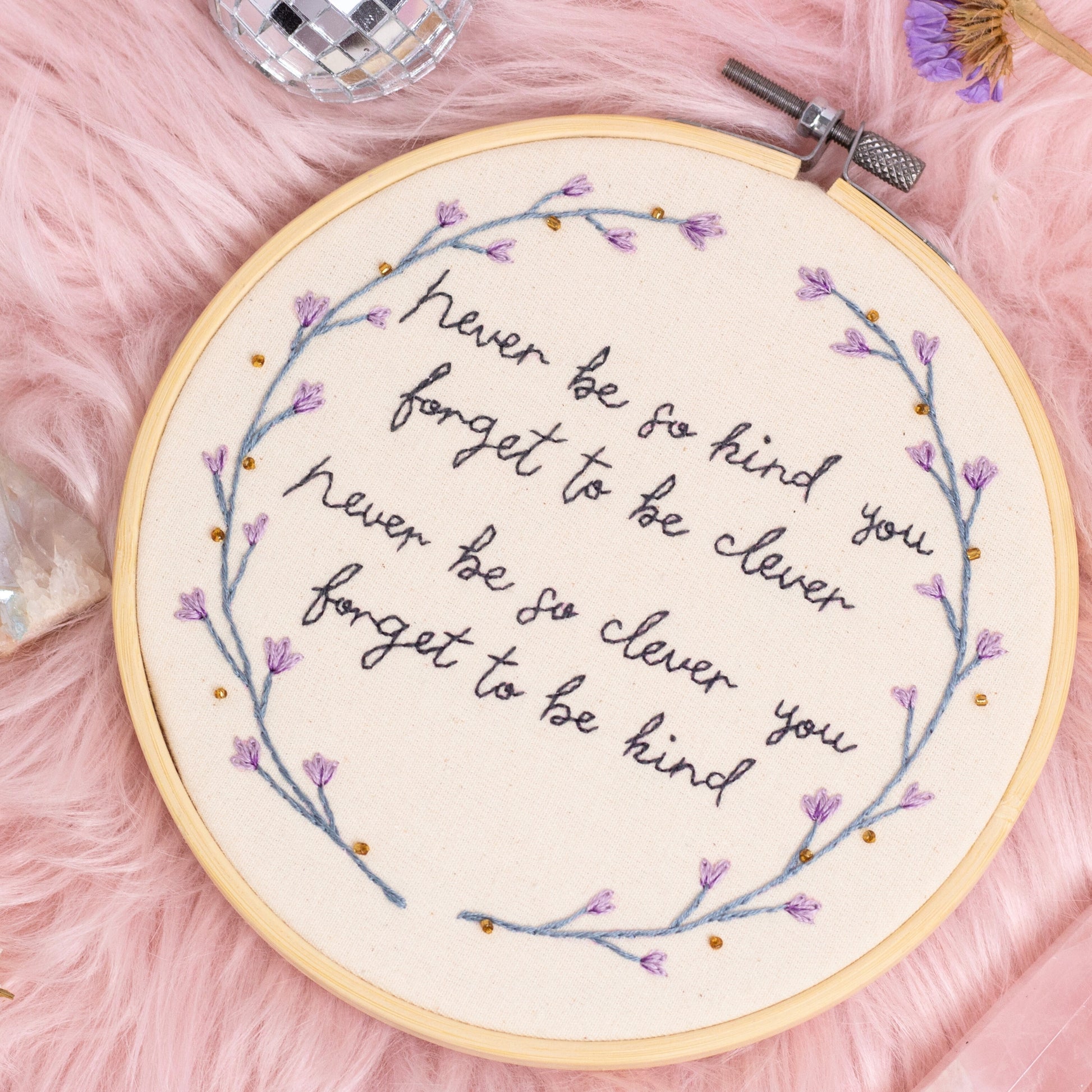Hand embroidered quote hoop reading never be so kind you forget to be clever surrounded by delicate purple floral wreath