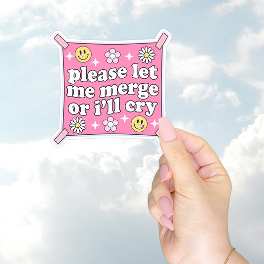 A hand holding a square pink bumper sticker styled like a flag that reads "please let me merge or I’ll cry" with daisies and smiley faces.