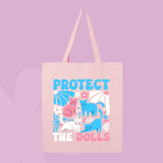 Pink tote bag with bold text “Protect the Dolls” and colorful illustrations of pink, blue, and white cats surrounded by flowers.