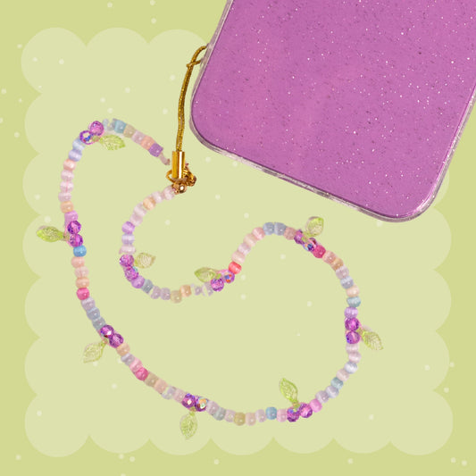 Purple cherry beaded phone wristlet with pastel rainbow beads, purple cherry charms, green leaf details, sparkly pink accents, and a gold phone strap attachment.