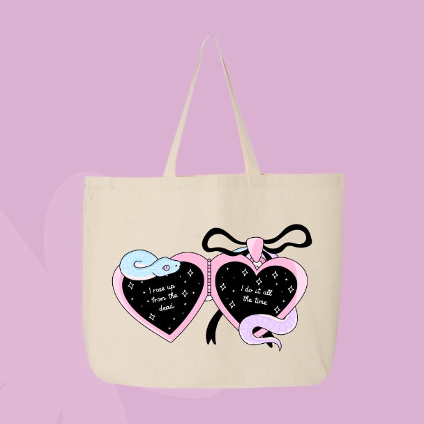 Beige tote bag with a locket design featuring pastel snakes and heart charms with the phrases “I rose up from the dead” and “I do it all the time.”