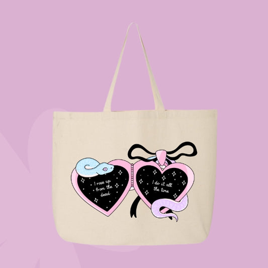 Beige tote bag with a locket design featuring pastel snakes and heart charms with the phrases “I rose up from the dead” and “I do it all the time.”