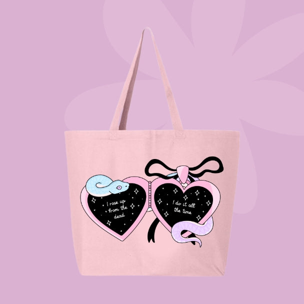 Pink tote bag with a locket design featuring pastel snakes and heart charms with the phrases “I rose up from the dead” and “I do it all the time.”