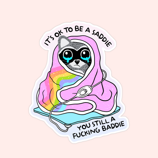 Sticker of a raccoon wrapped in a rainbow blanket with the phrase "It’s ok to be a saddie, you still a f***ing baddie."