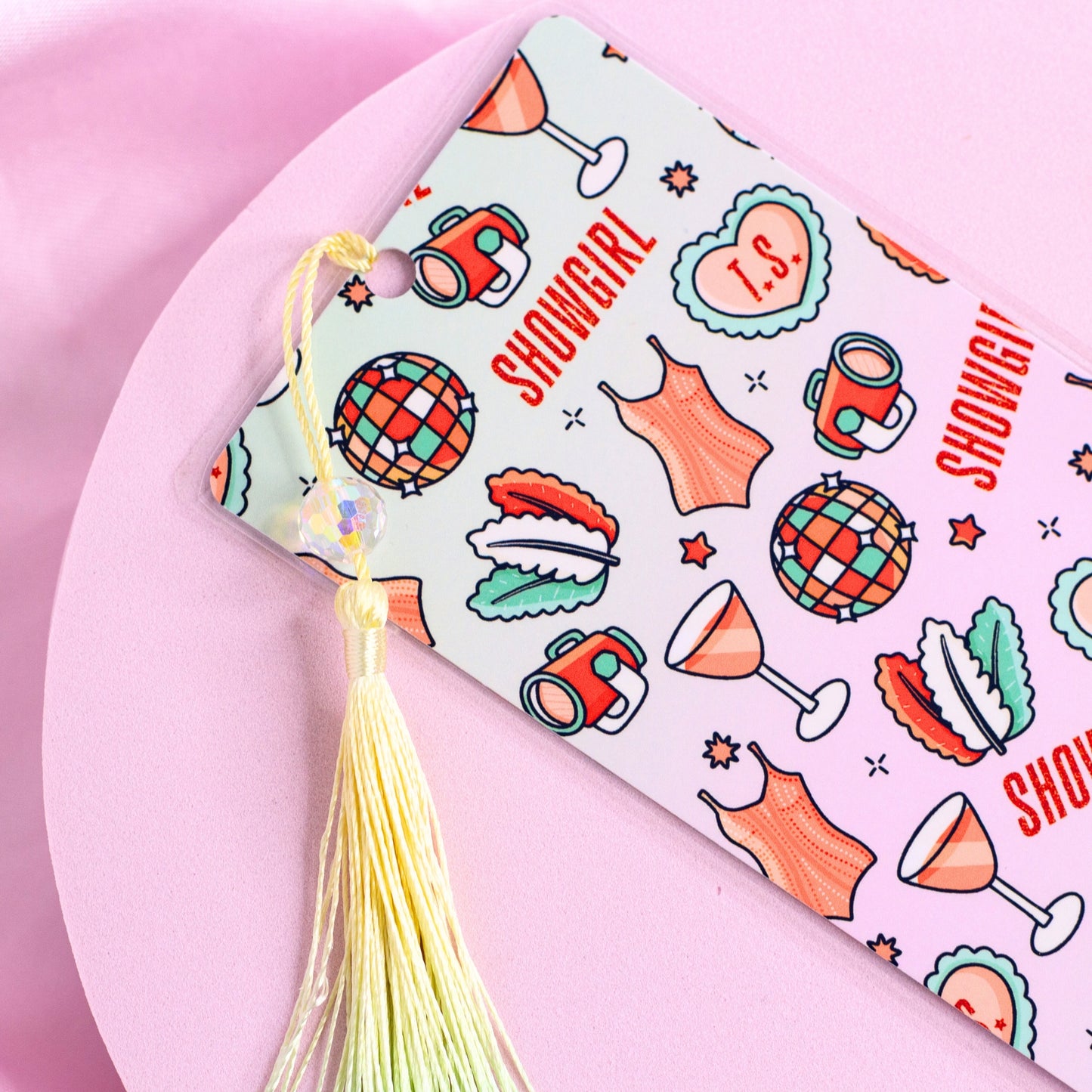 White bookmark with repeating disco balls, cocktails, corsets, and “Showgirl” text, finished with a pastel tassel close up.